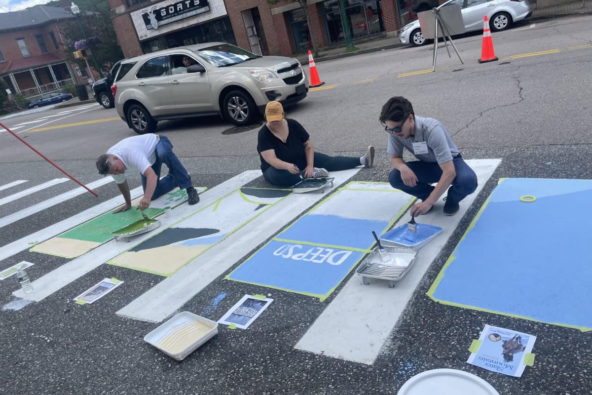 Three people are painting colorful designs on a crosswalk in global city Norwich while sitting on the street, with paint supplies and reference images nearby. A car and orange cones are seen in the background.
