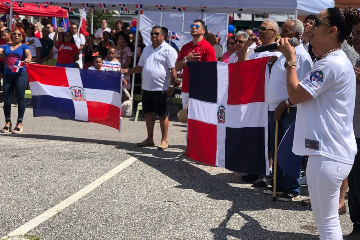 A group of people stand outdoors in Global City Norwich, holding Dominican Republic flags while a woman sings or speaks into a microphone.