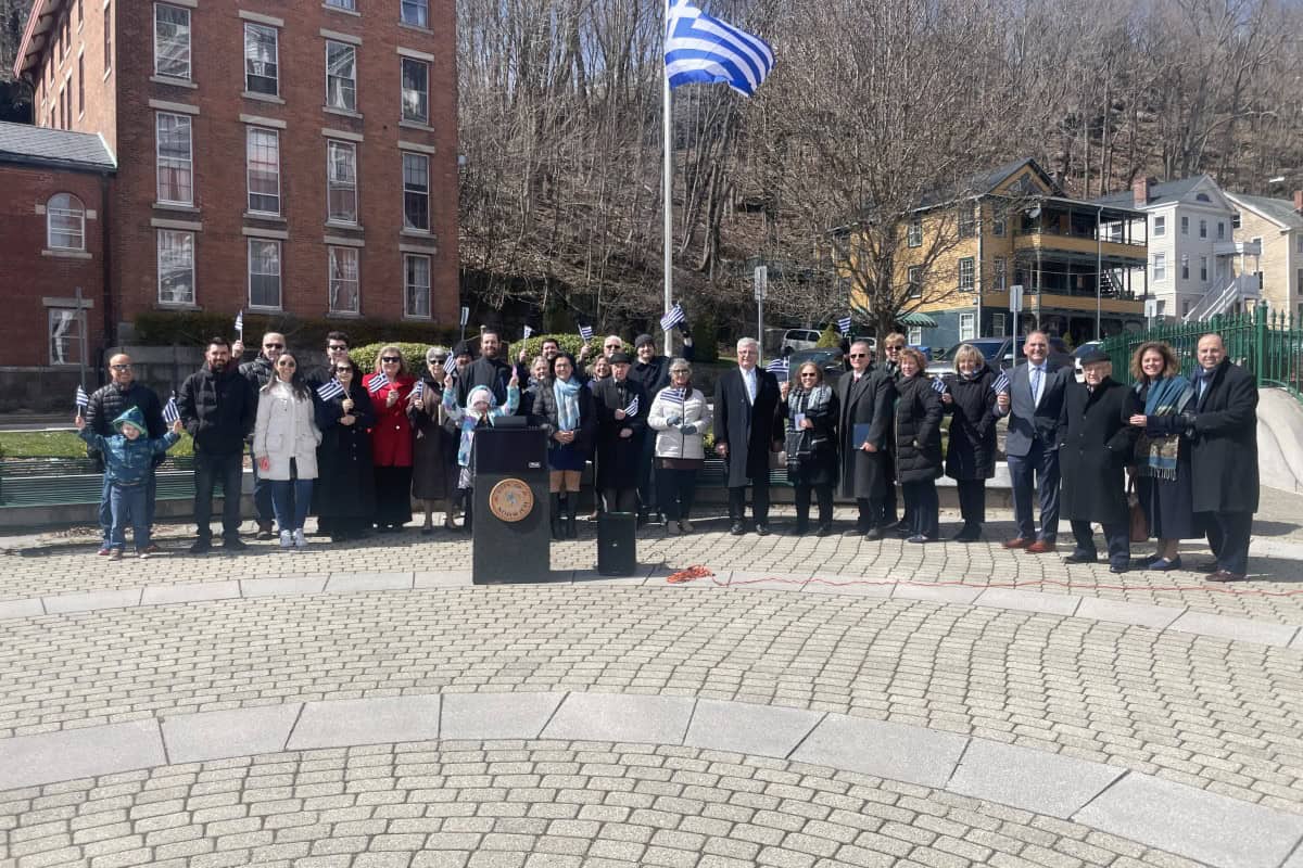 A group of people stands together outdoors near a podium and a Greek flag in global city Norwich, with buildings and trees in the background.