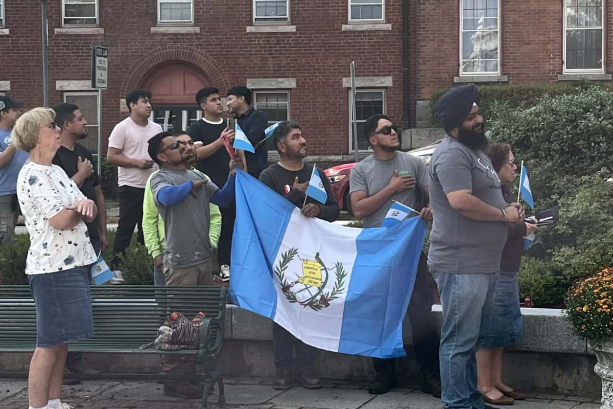 A group of people stand outdoors in global city Norwich, holding and waving Guatemalan flags, some with hands on their chests, in front of a brick building.