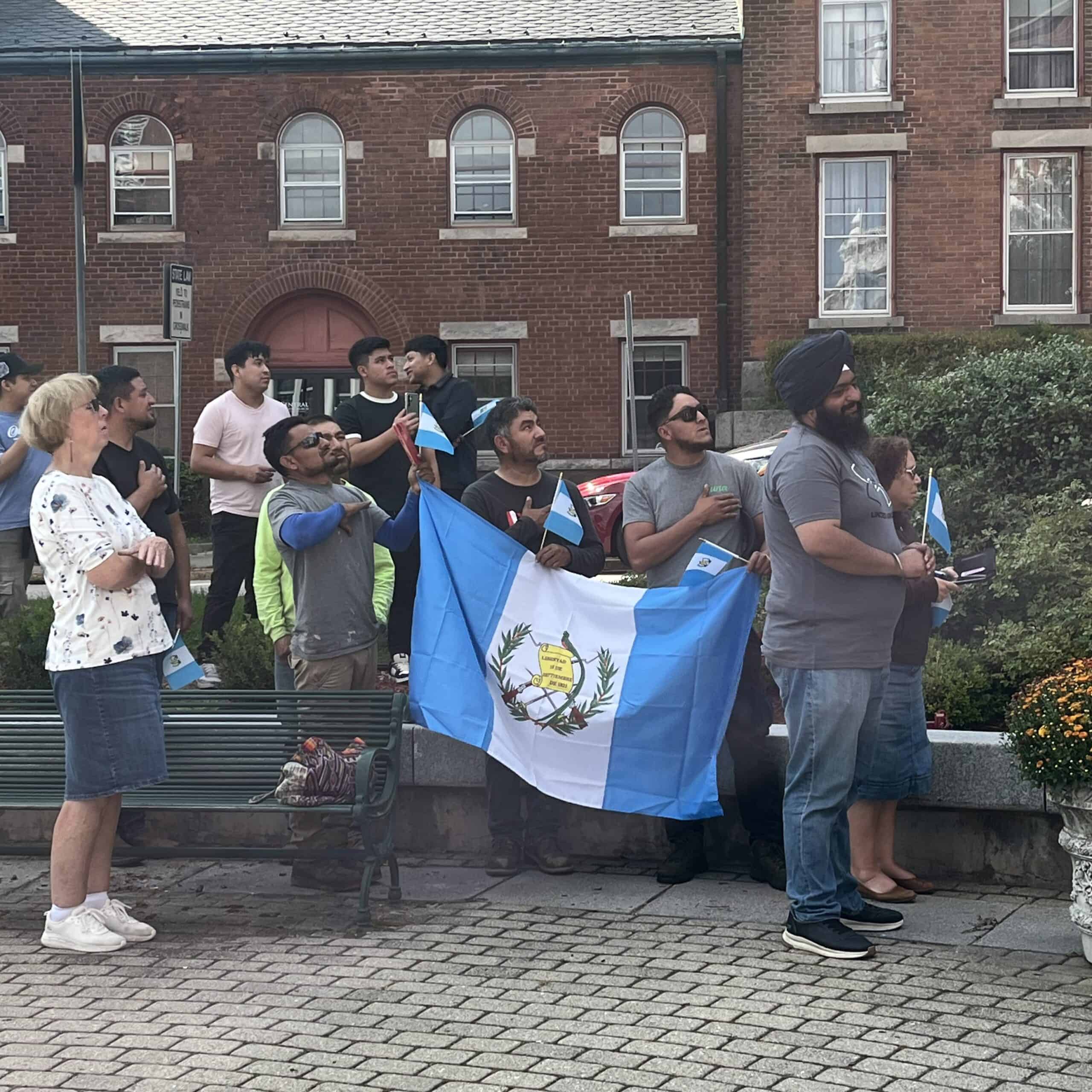 A group of people stands outdoors in global city Norwich, holding small Guatemalan flags and a large Guatemalan flag, with hands over their hearts in front of brick buildings.