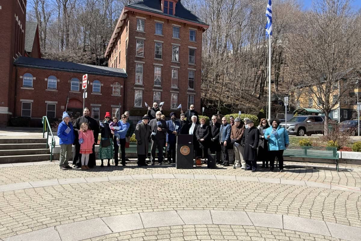 A group of people stands around a podium in an outdoor plaza in global city Norwich, with a brick building and flagpole in the background on a sunny day.