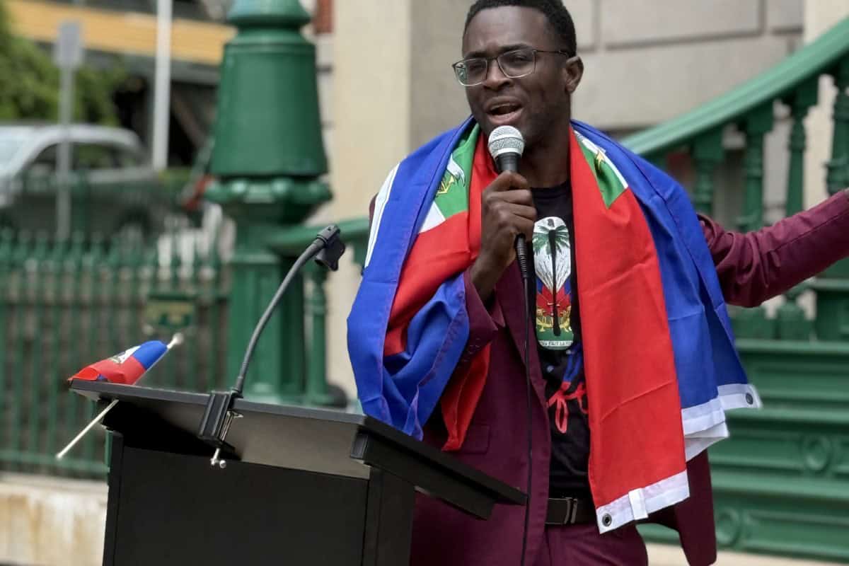 A man wearing glasses and a flag draped over his shoulders speaks into a microphone at an outdoor podium in global city Norwich.