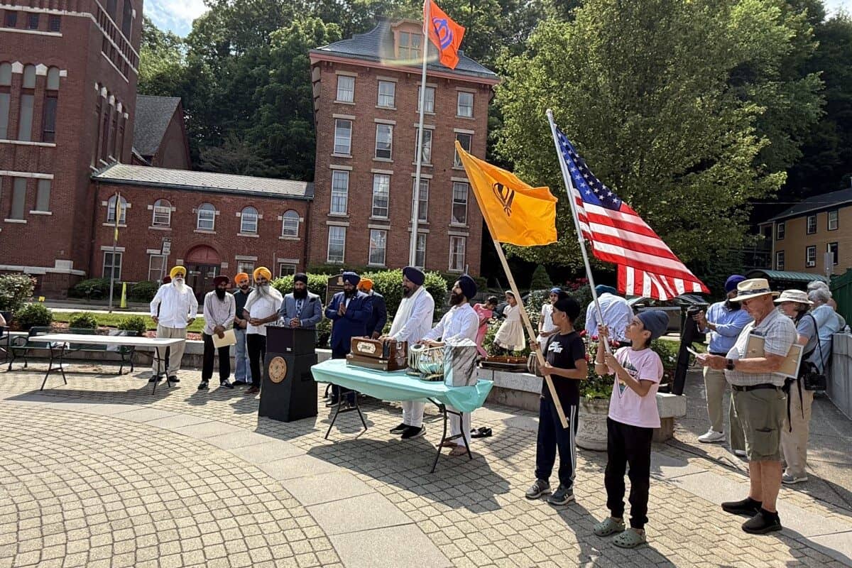 A group of people stand outdoors near a podium with American and orange flags in global city Norwich; some wear turbans, and various items are displayed on tables. Brick buildings and trees form the backdrop.