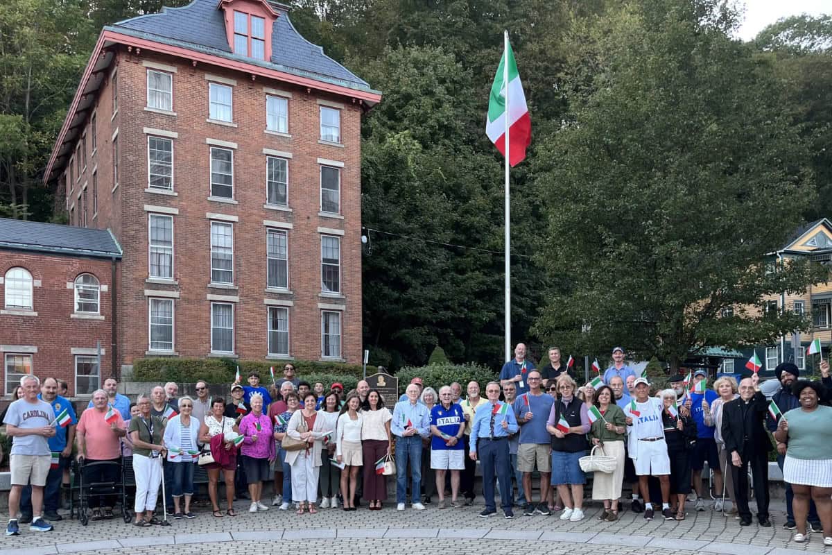 A large group of people stands together outdoors near a flagpole with the Italian flag, in front of a historic brick building and trees, capturing the vibrant spirit of global city Norwich.