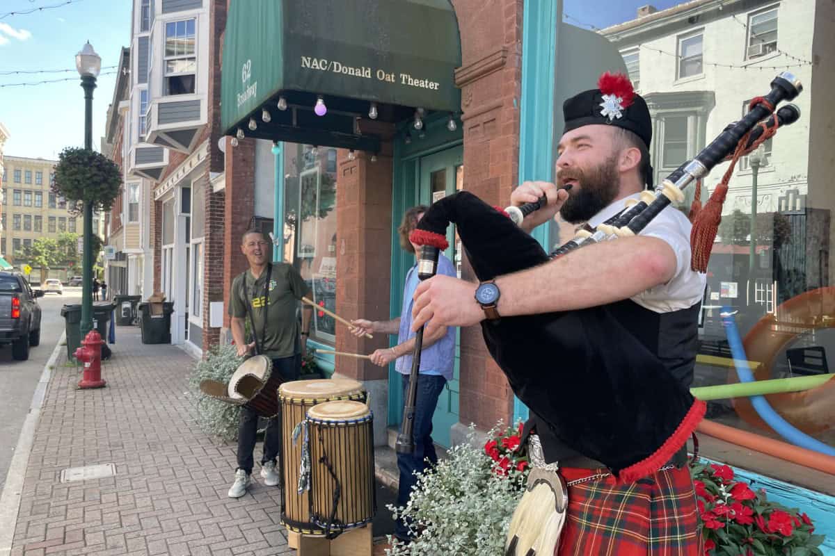 A man in a kilt plays bagpipes on a sidewalk while two others play drums outside the NAC/Donald Oat Theater in global city Norwich on a sunny day.