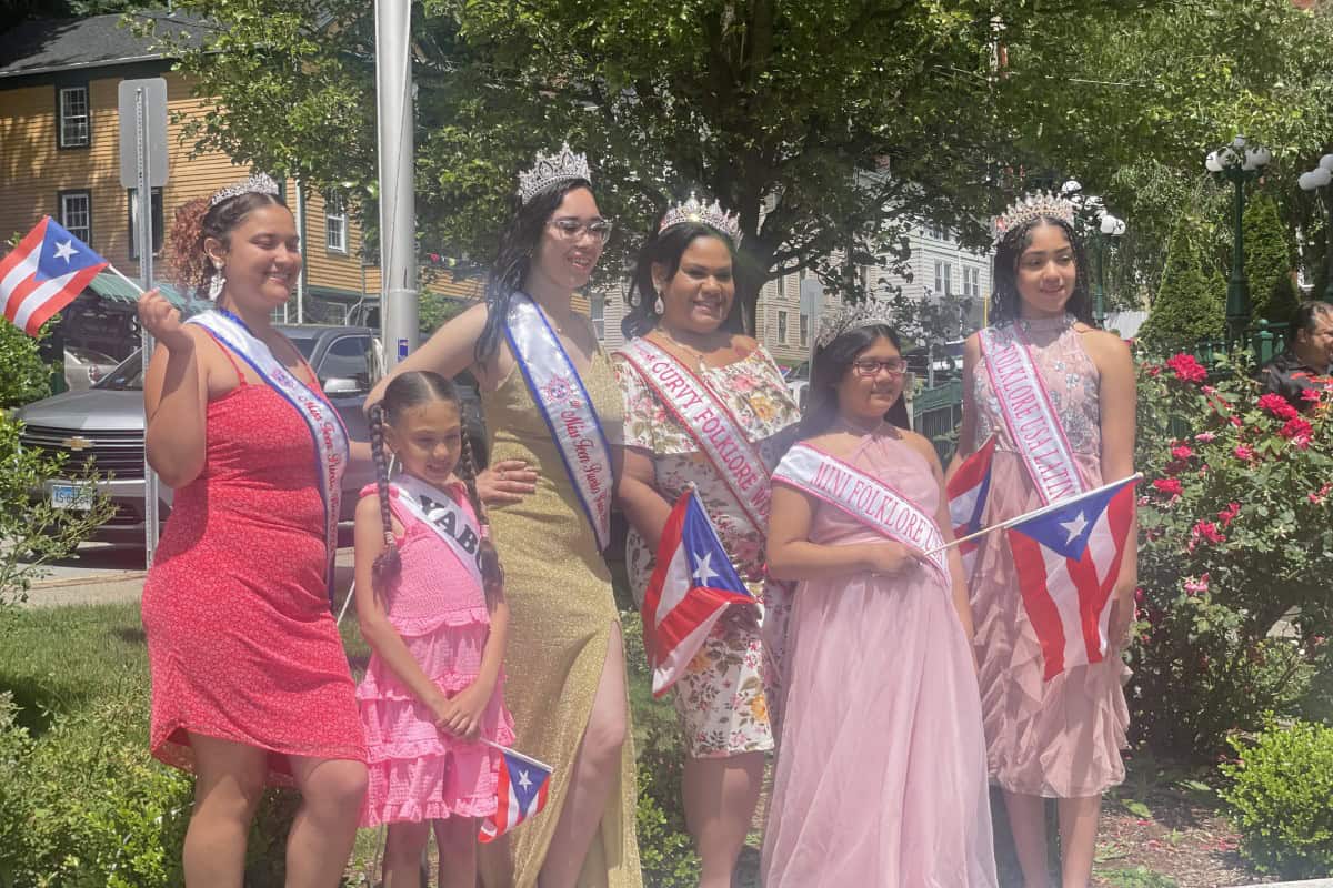 Six women and girls wearing sashes and crowns stand outdoors in global city Norwich, holding Puerto Rican flags, posing together in sunny weather near flowers and greenery.