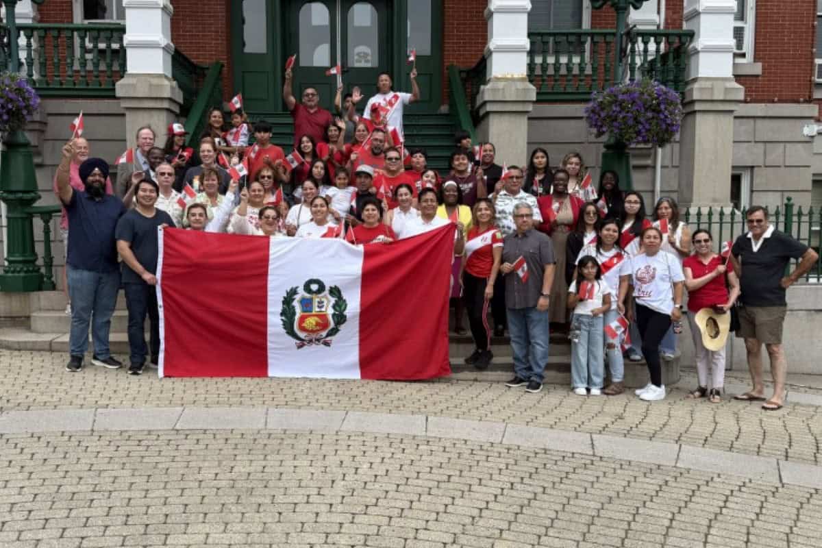 A large group of people, many dressed in red and white, pose outdoors holding a Peruvian flag and small flags in front of a building with steps and hanging flower baskets in global city Norwich.