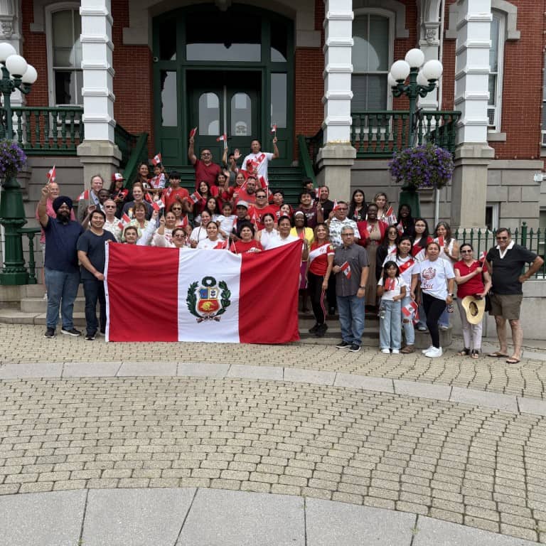 A large group of people standing in front of a building in global city Norwich, holding a Peruvian flag and wearing red and white clothing.