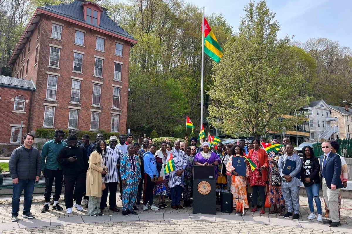 A group of people stand outdoors near a flagpole displaying the Togo flag in global city Norwich, with several holding smaller flags, in front of a brick building and trees.