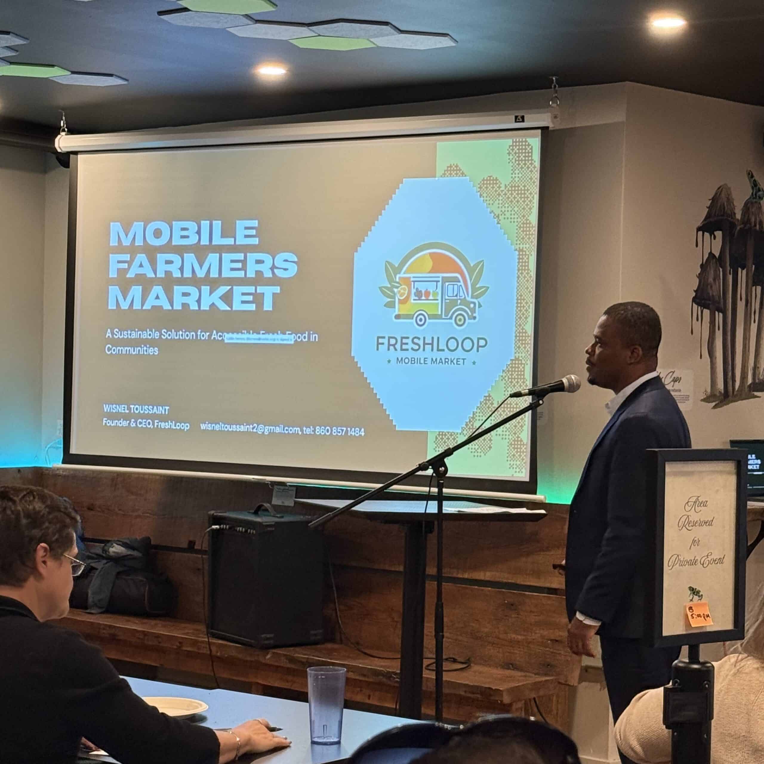 A man stands at a lectern giving a presentation about "Mobile Farmers Market" and "FreshLoop" at an indoor event in global city Norwich, with a projected slide displayed behind him.